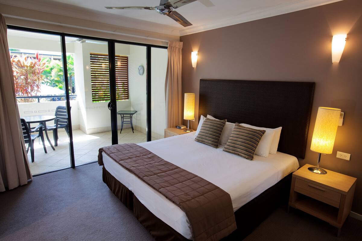 A hotel room with a large bed, two bedside tables with lamps, a ceiling fan, and a sliding glass door leading to an outdoor area with a table and chairs.