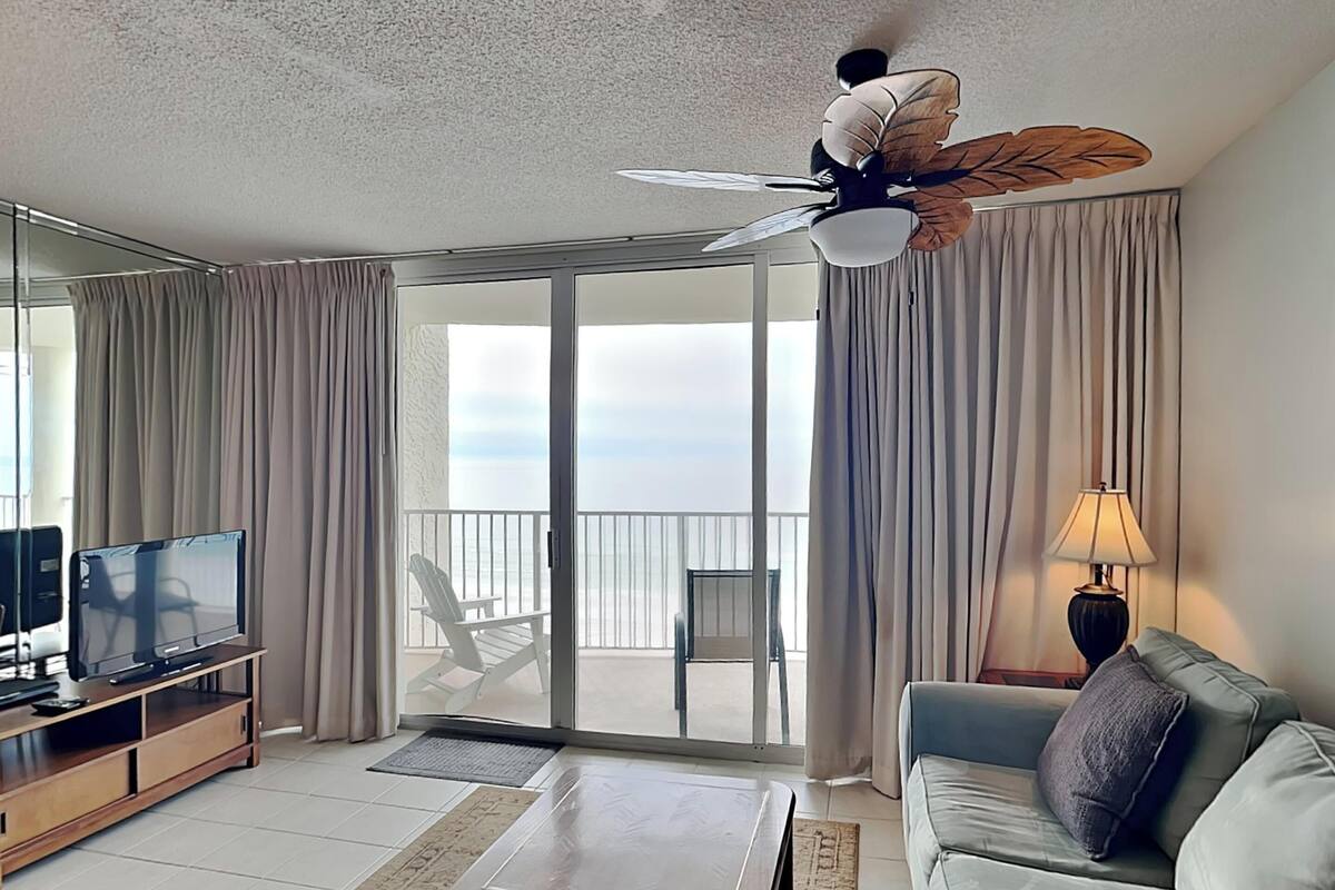 A modern hotel room with a large sliding glass door leading to a balcony, a flat-screen TV, a sofa, a coffee table, and a ceiling fan with a leaf design.