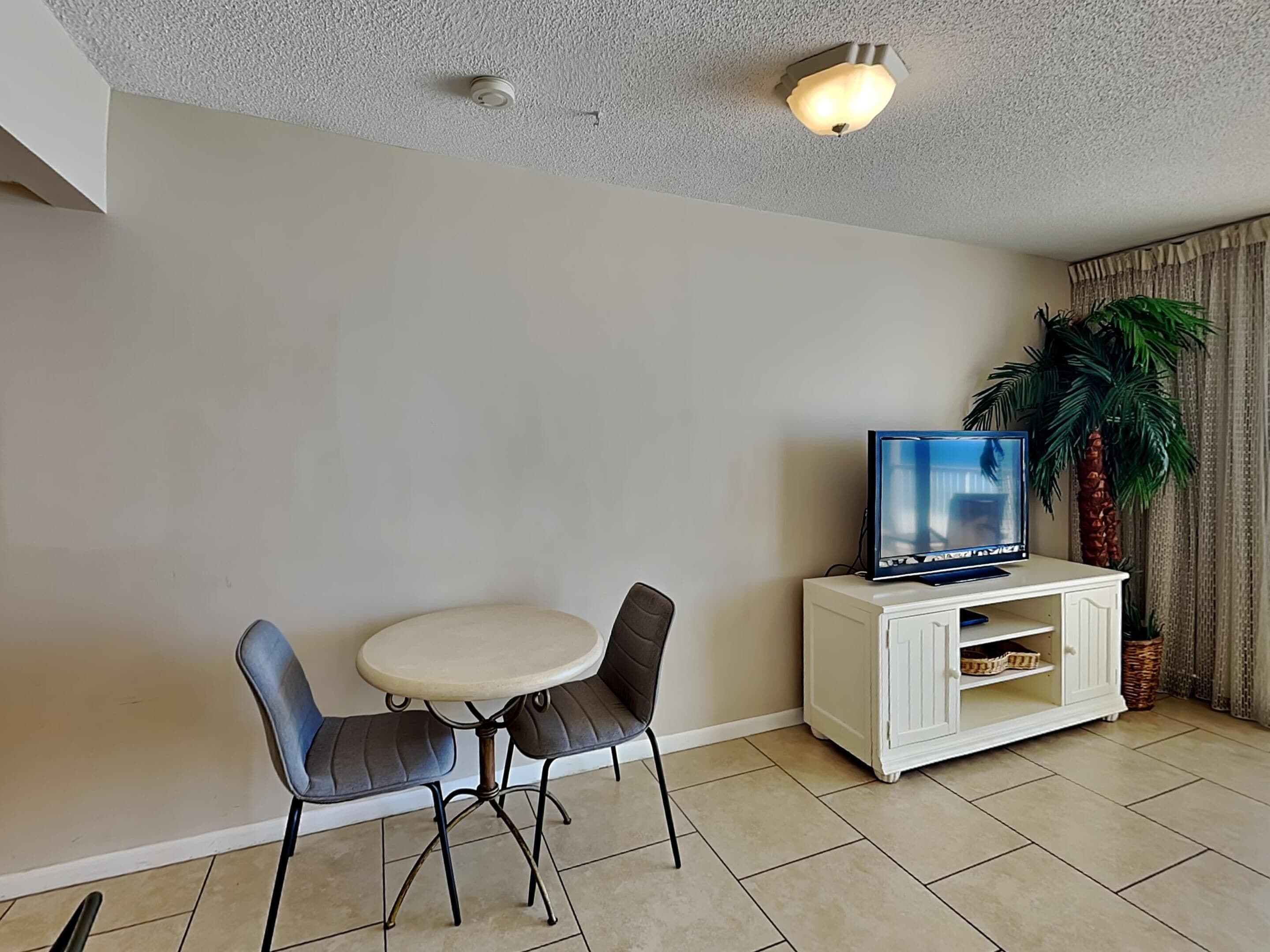 A living room with a TV on a white cabinet, a round table with two chairs, a ceiling light, and a potted plant.