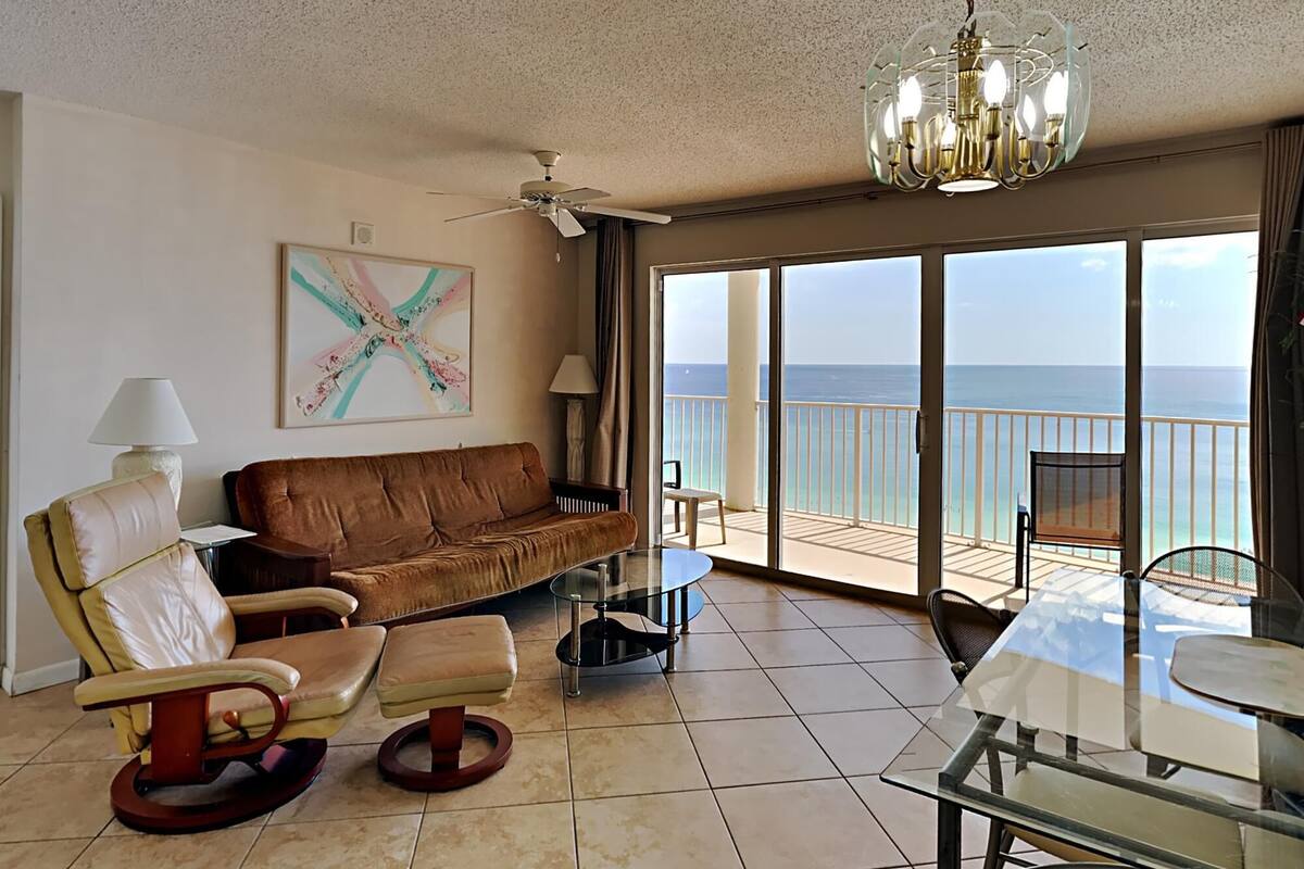 A living room with a brown sofa, a glass table, and a view of the ocean through large sliding doors.