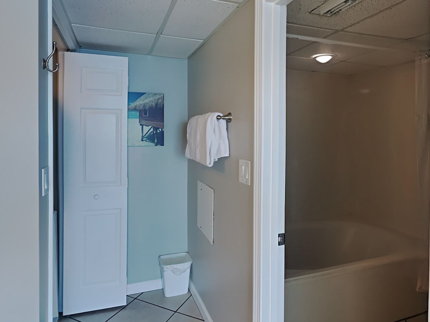 A bathroom with a white door, a small trash bin, a towel hanging on a hook, and a picture on the wall.