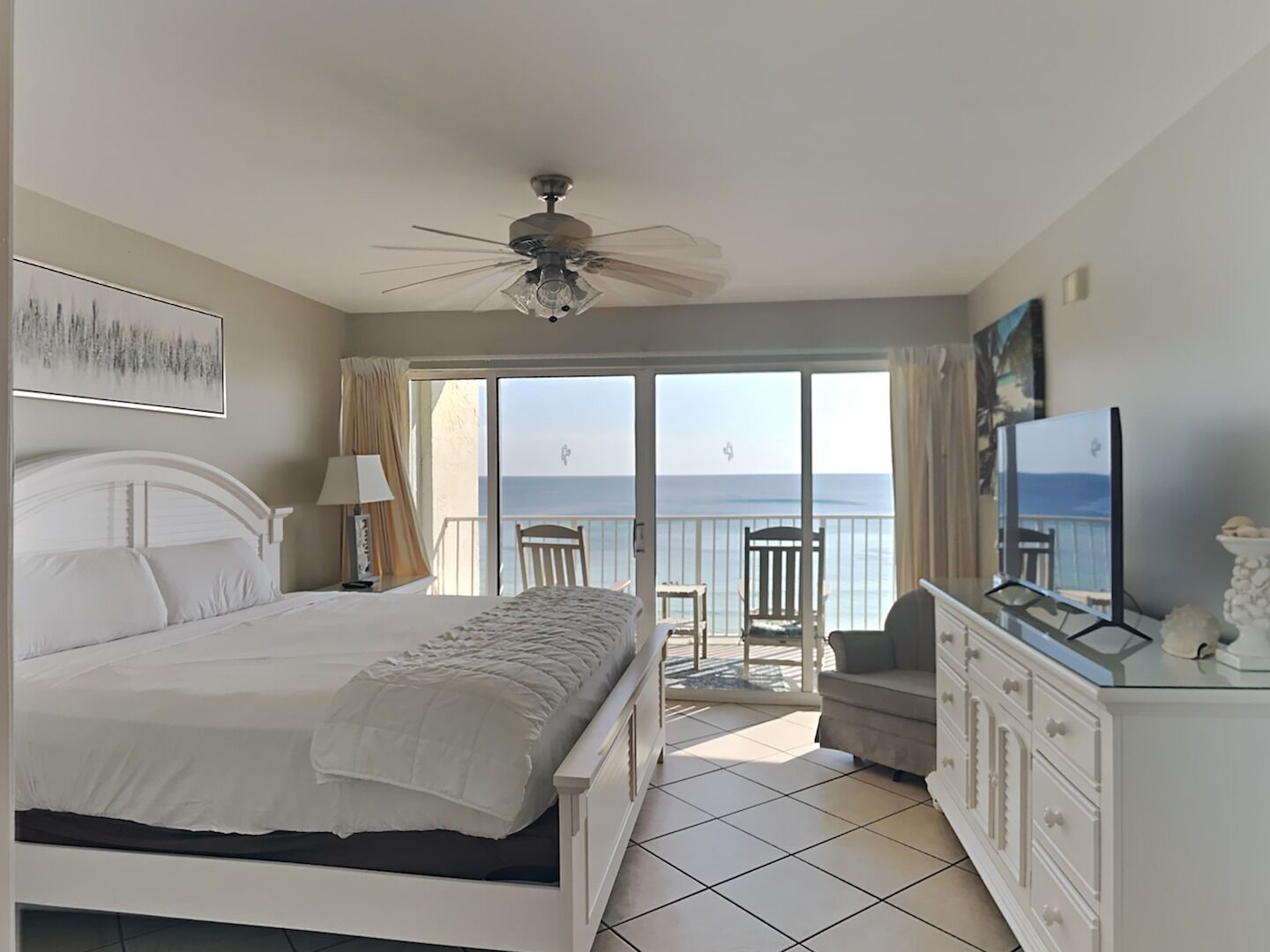 A bedroom with a bed, a ceiling fan, a TV, and a balcony with a view of the sea.