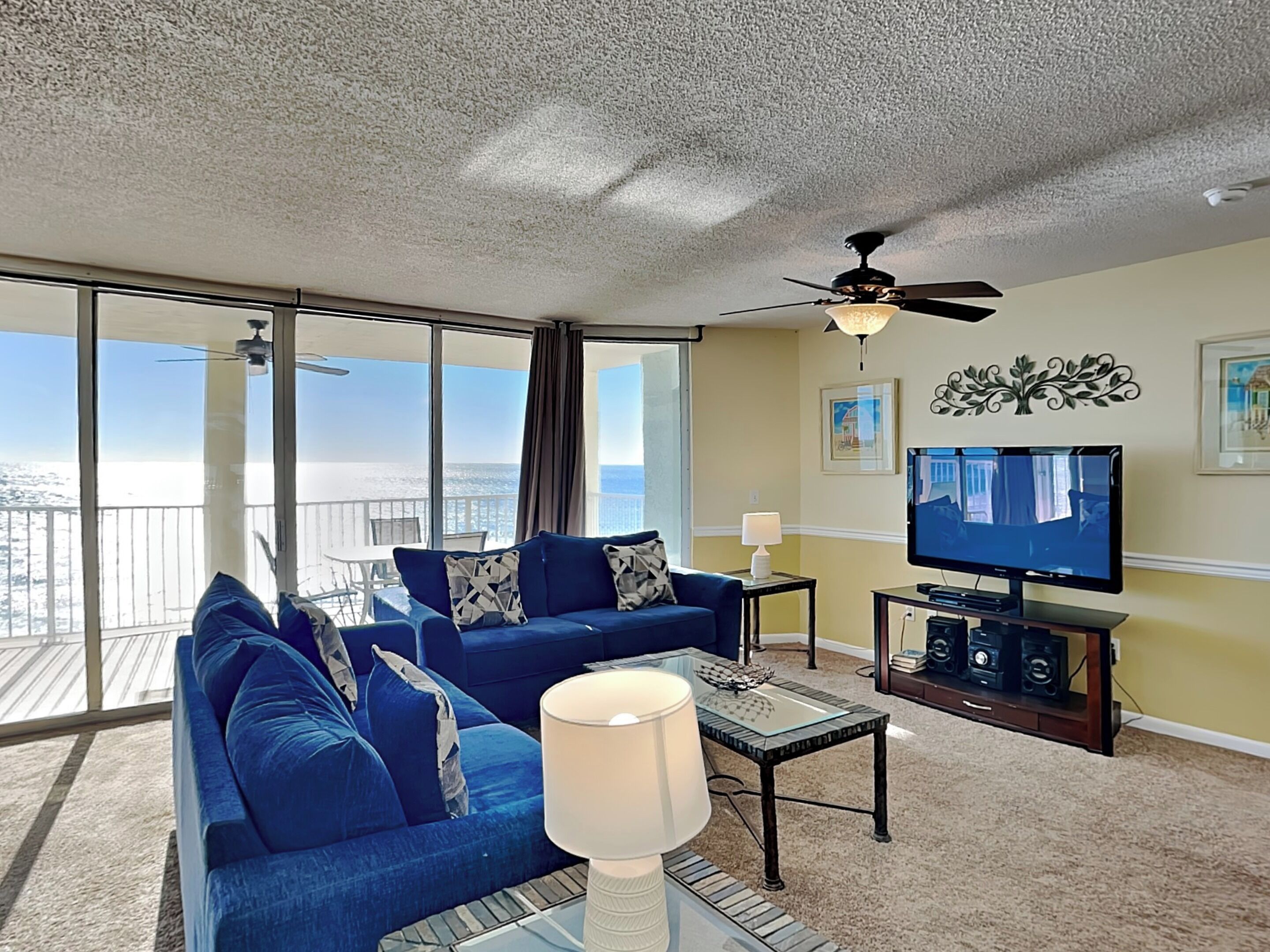 Een ruime woonkamer met een blauwe bank, een salontafel, een televisie en een balkon met uitzicht op de oceaan.