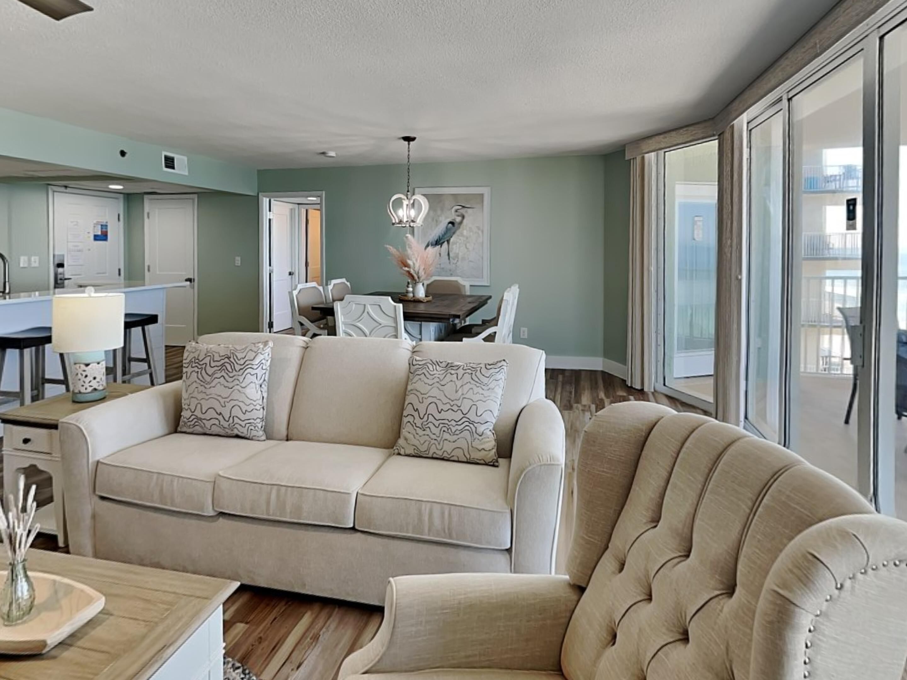 Una sala de estar con un sofá beige, una zona de comedor con mesa y sillas, y ventanas amplias que ofrecen una vista.