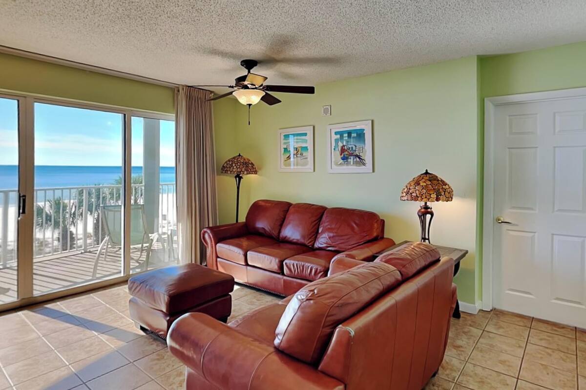 A spacious living room with a brown leather sofa, a ceiling fan, and a view of the ocean through large sliding glass doors.
