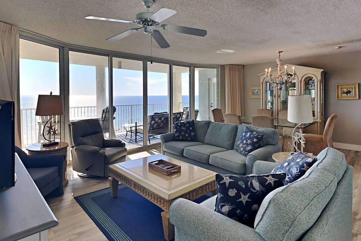 A spacious living room with a large window overlooking the ocean, a ceiling fan, and a chandelier.