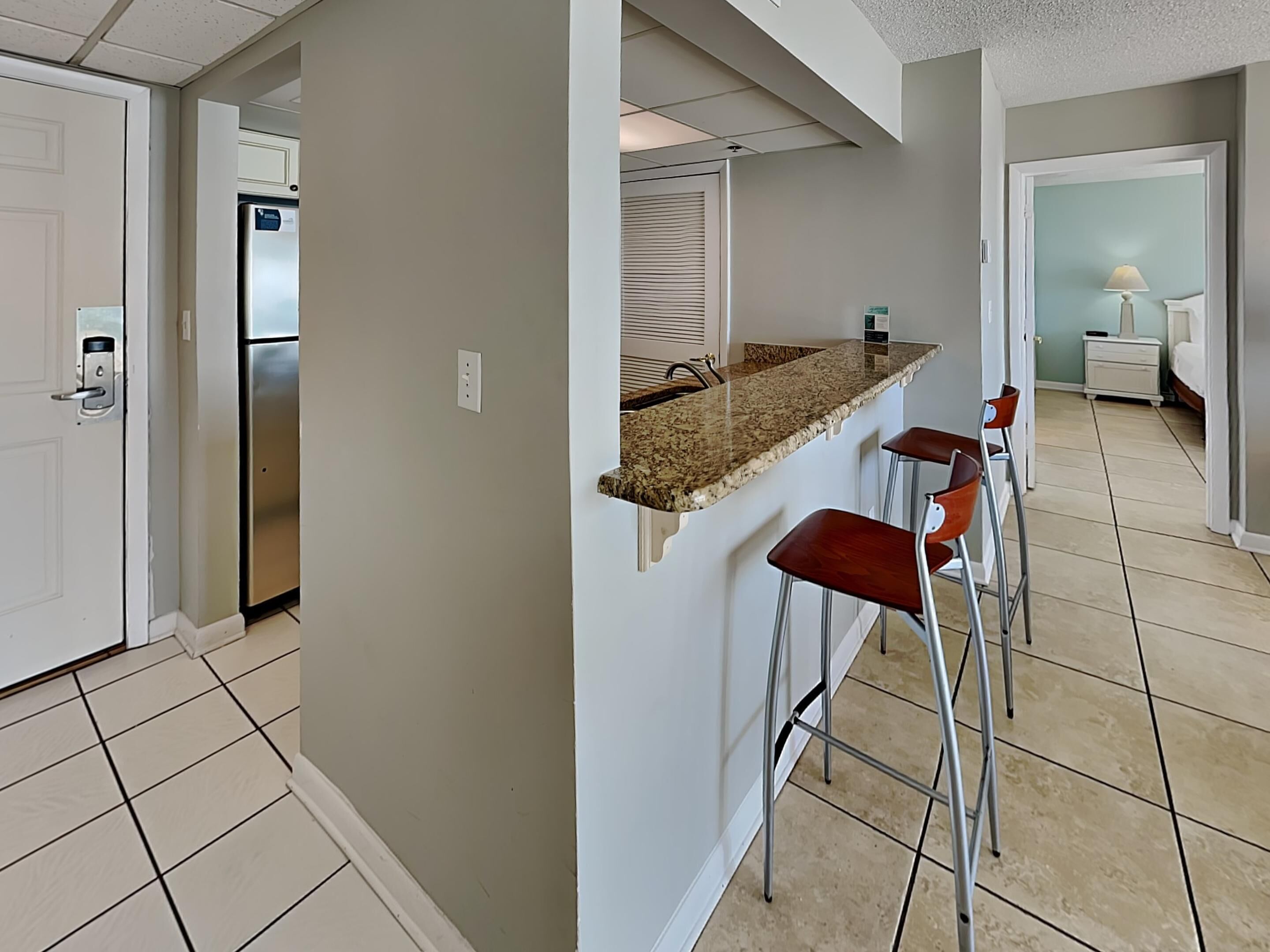 A modern kitchen with a granite countertop, stainless steel refrigerator, and a built-in oven. There are two bar stools and a window with blinds.