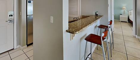 A modern kitchen with a granite countertop, stainless steel refrigerator, and a built-in oven. There are two bar stools and a window with blinds.
