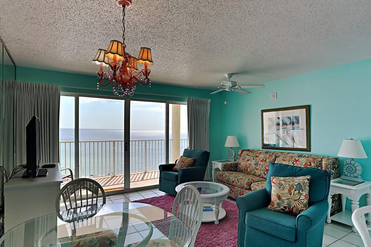 A living room with a view of the ocean, a chandelier, teal sofas, and a ceiling fan.