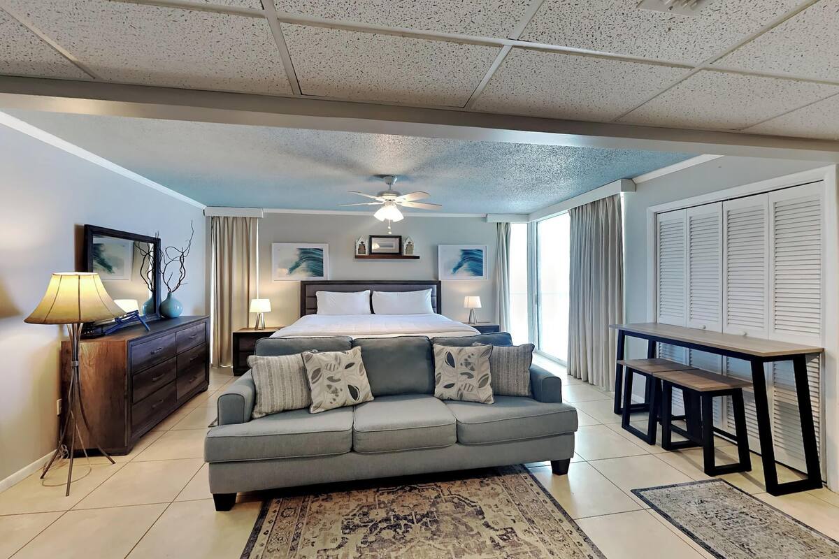 A modern hotel room with a large bed, a grey sofa, a dining table, and a ceiling fan.