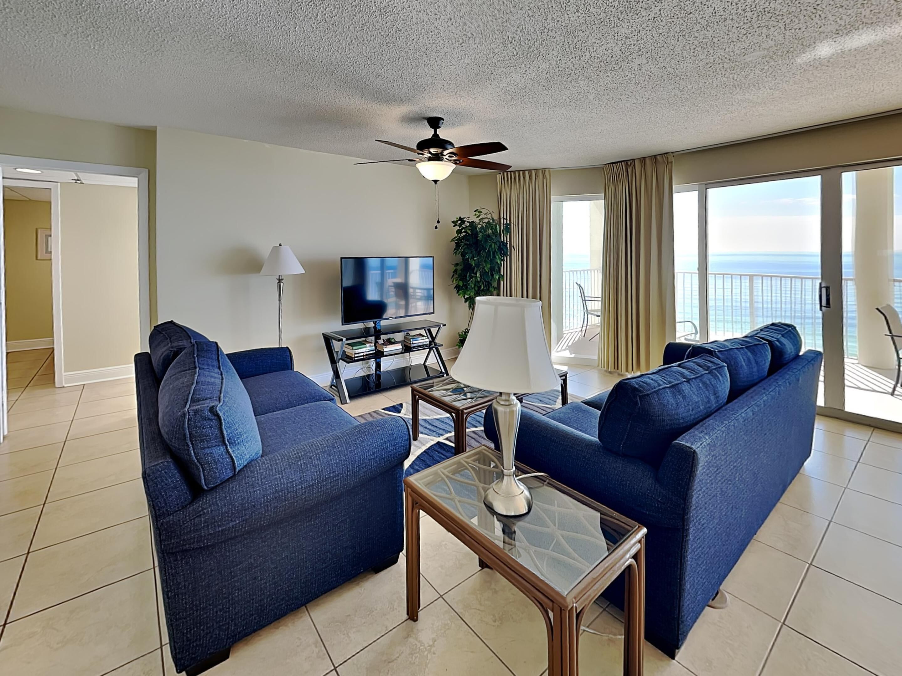A living room with two blue sofas, a glass coffee table, a TV on a stand, and a ceiling fan.