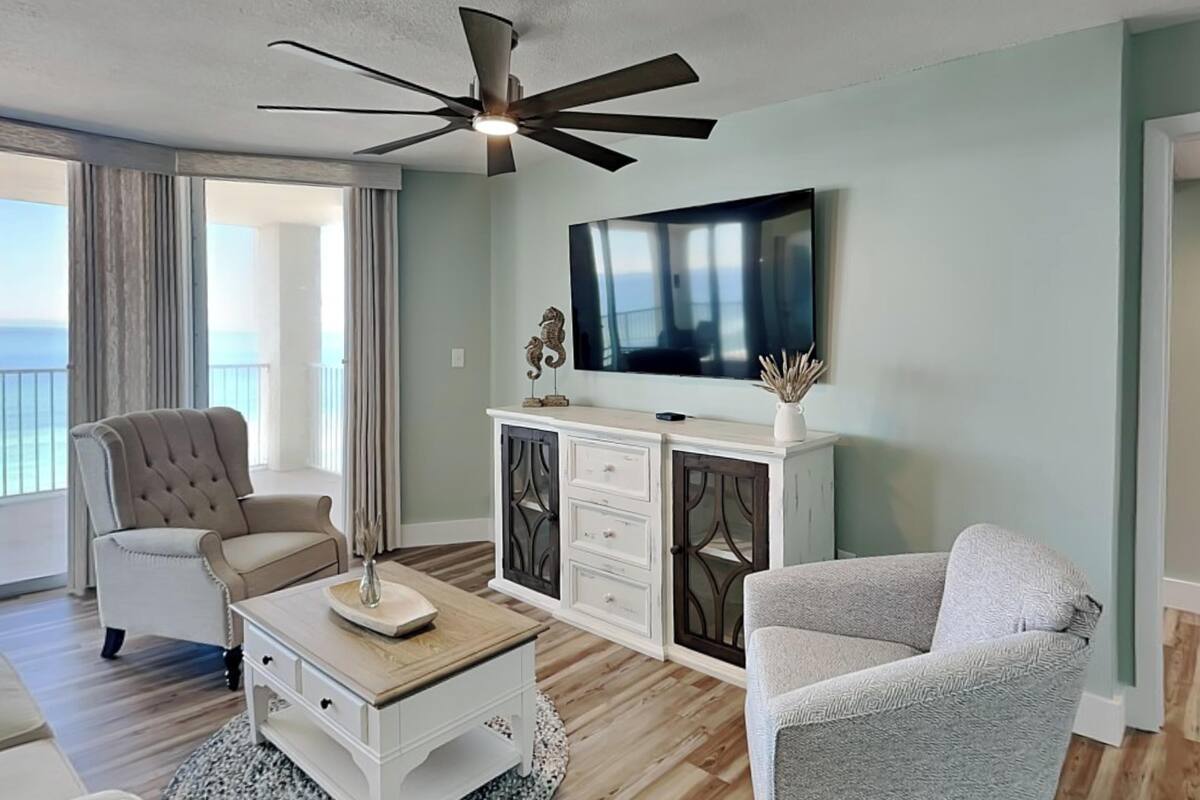 A living room with a ceiling fan, a flat-screen TV, a beige armchair, a white cabinet, and a grey armchair.