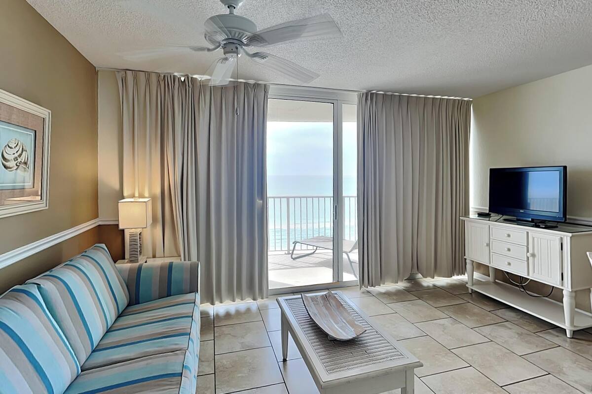A hotel room with a striped sofa, a small table, a television on a cabinet, and a balcony with curtains.