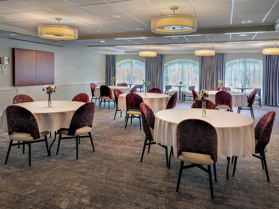 Un salón de conferencias con mesas y sillas redondas, ventanas amplias con cortinas y un diseño de techo moderno.
