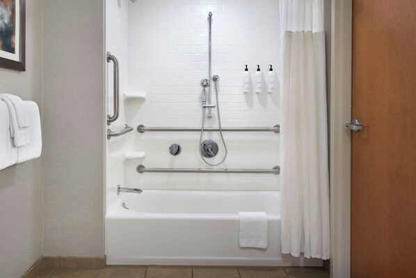 Un baño de hotel estándar con bañera, barras de apoyo, cabezal de ducha y un perchero para toallas.
