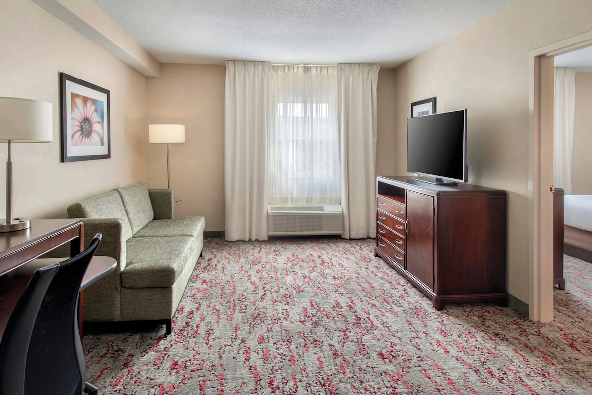 Habitación de hotel con un sofá verde, un televisor de pantalla plana sobre un soporte de madera, un escritorio con una silla y una ventana grande con cortinas translúcidas.