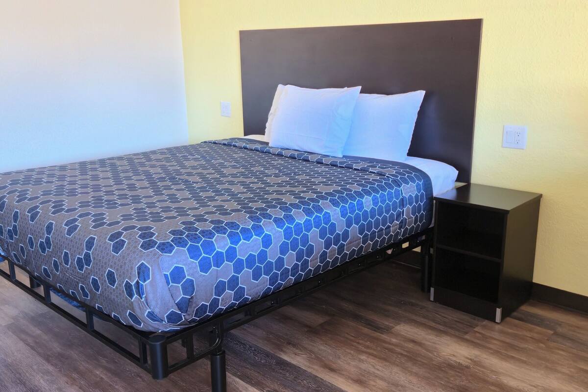 A bed with a patterned bedspread, a headboard, white pillows, a nightstand, and a wooden floor.