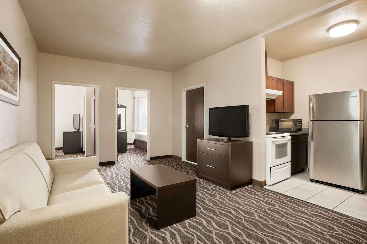 Habitación de hotel con sofá, televisor, una pequeña cocina equipada con refrigerador y microondas, y alfombra estampada.