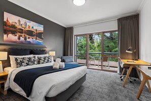 Une chambre d’hôtel avec un grand lit, un bureau, une chaise, un balcon donnant sur un paysage verdoyant et un tableau représentant une vue sur la ville.