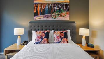 Una habitación de hotel con una cama, dos mesitas de noche, un cuadro de una bicicleta en un muelle y una lámpara de noche en cada mesita.