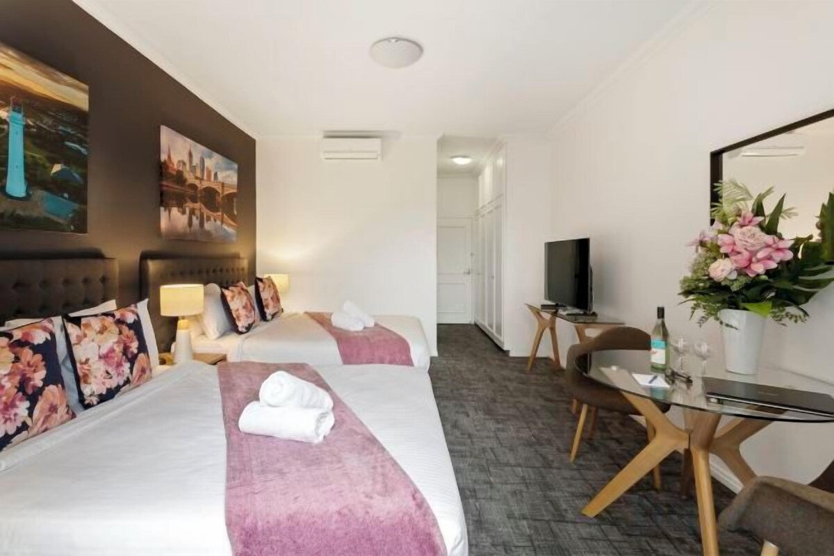 Une chambre d’hôtel avec deux lits, une télévision, un bureau avec un ordinateur et une salle de bain visible au fond.