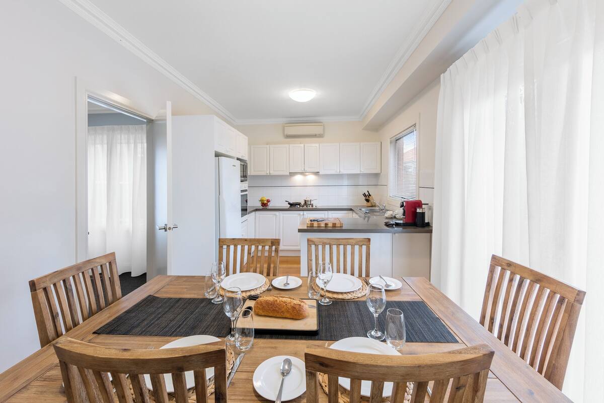 Un coin repas avec une table en bois disposée pour quatre personnes, entourée de chaises en bois, dans une cuisine moderne avec des meubles blancs et un four encastré.