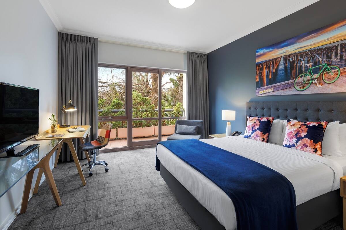 Une chambre d’hôtel avec un grand lit, un bureau avec un ordinateur, une chaise, une télévision et un balcon donnant sur un paysage verdoyant.