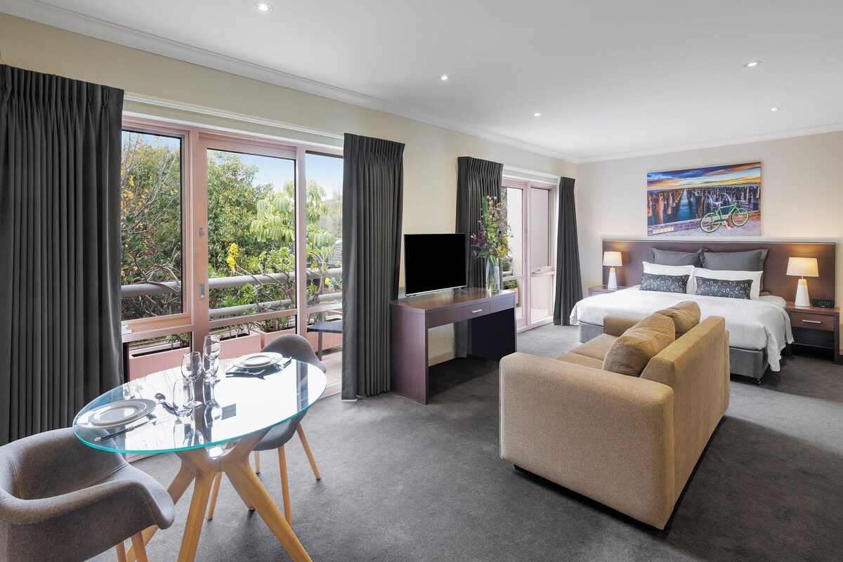 Ein Hotelzimmer mit Esstisch aus Glas, einer Couch, einem Bett, einem Fernseher und einem Gemälde.