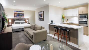 A modern hotel room with a bed, sofa, dining table, and kitchenette.