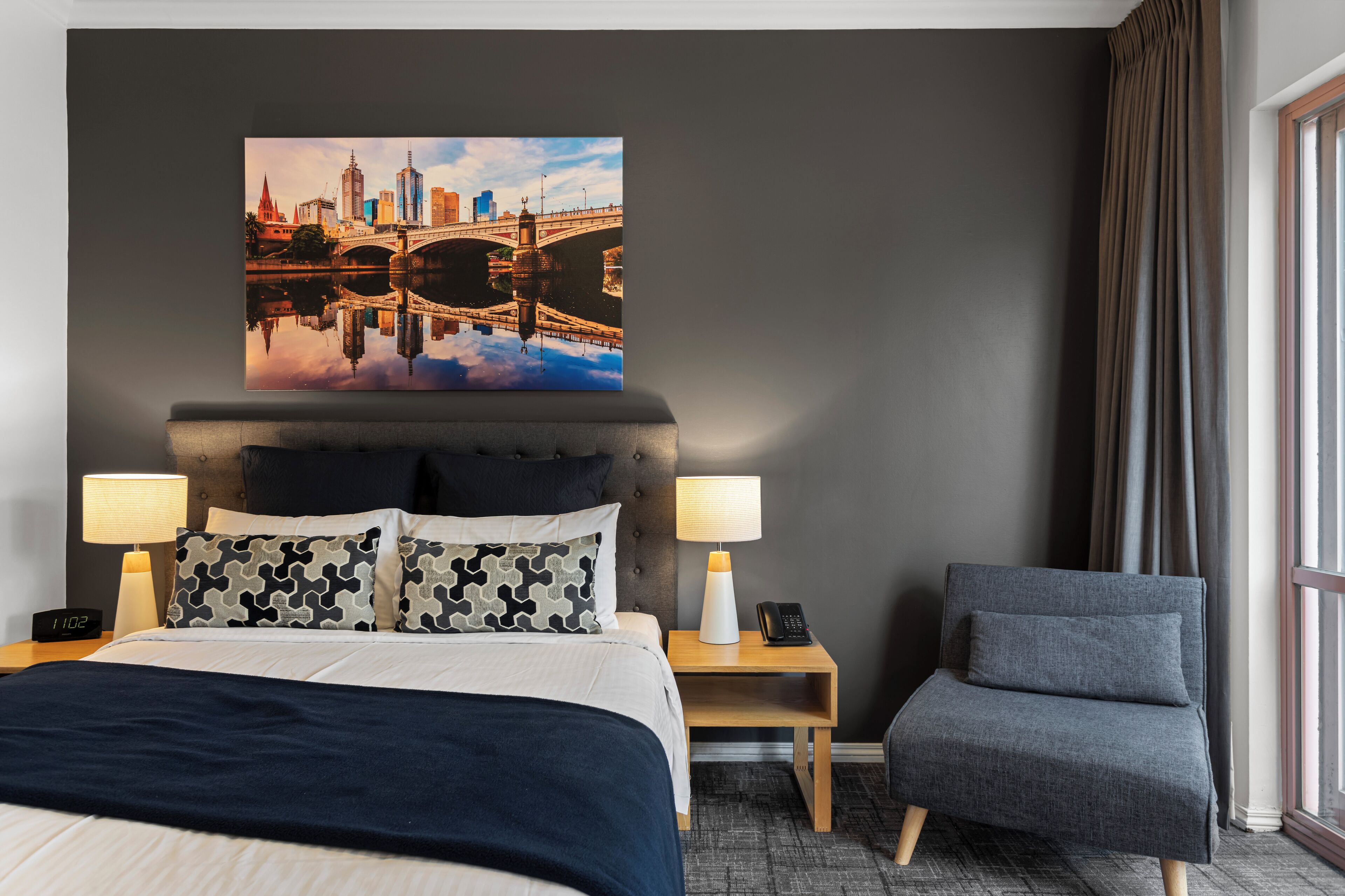 Habitación de hotel con cama, dos mesitas de noche, una silla, un cuadro grande de un paisaje urbano en la pared y una ventana con cortinas.