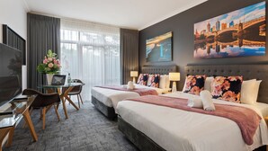 Quarto de hotel com duas camas, uma mesa com um laptop, uma televisão e uma janela grande com cortinas.