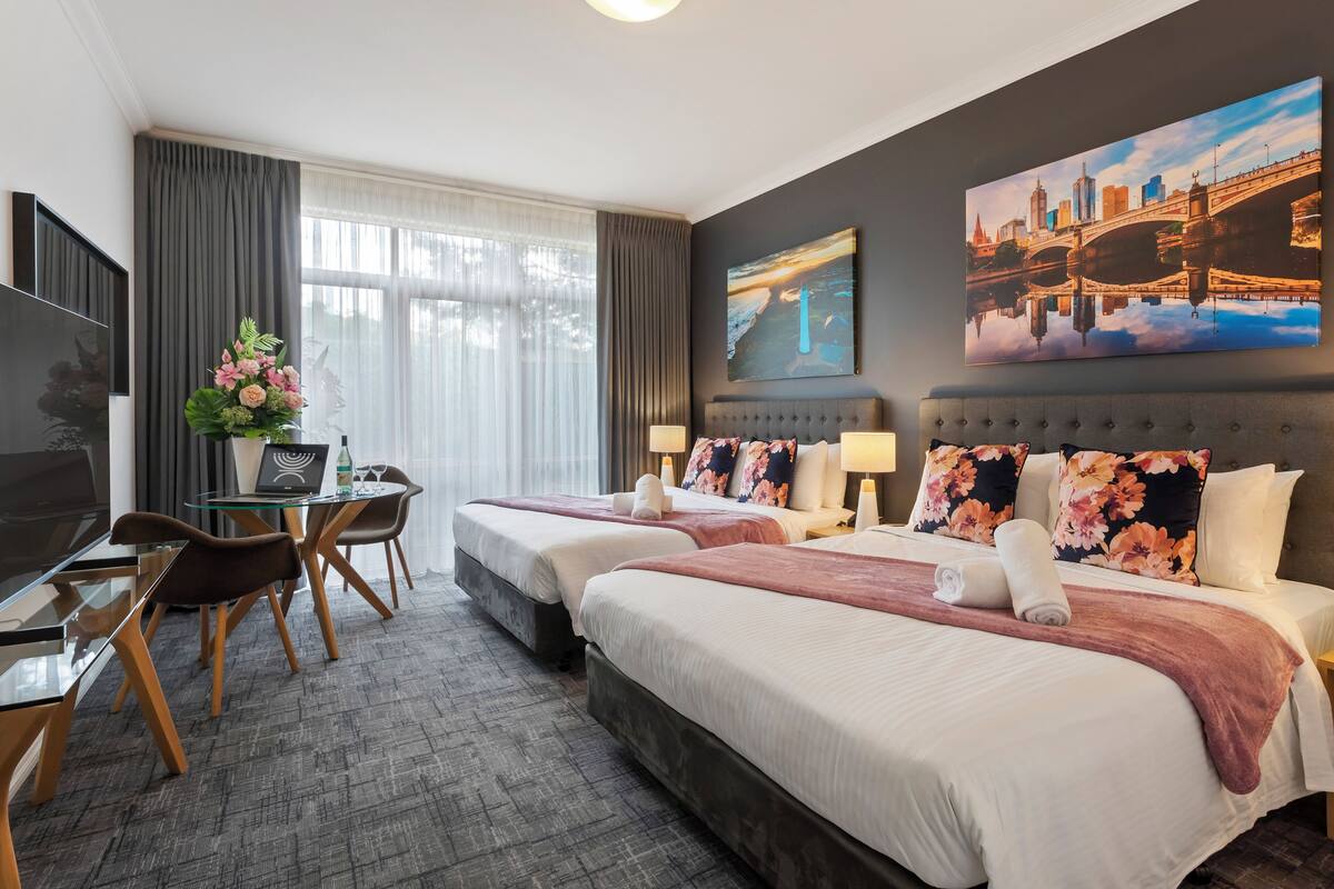 Une chambre d’hôtel avec deux lits, un bureau avec un ordinateur portable, une télévision et une grande fenêtre avec des rideaux.
