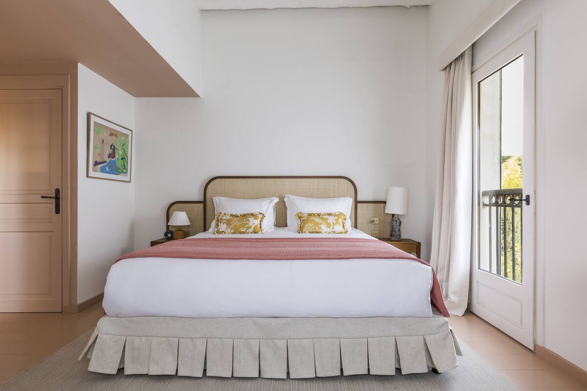 Een netjes opgemaakt bed met een wit-rode dekbedovertrek, twee witte kussens en een beige bedrok. Er staat een nachtkastje met een lamp bij, en links aan de muur hangt een schilderij.