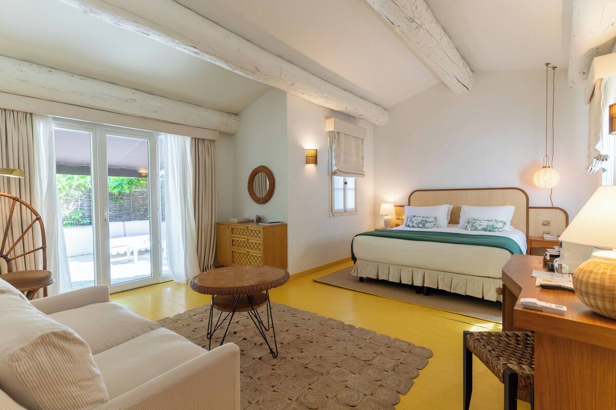 Een slaapkamer met een bed, een bureau, een stoel, een kleine tafel en een raam met gordijnen.