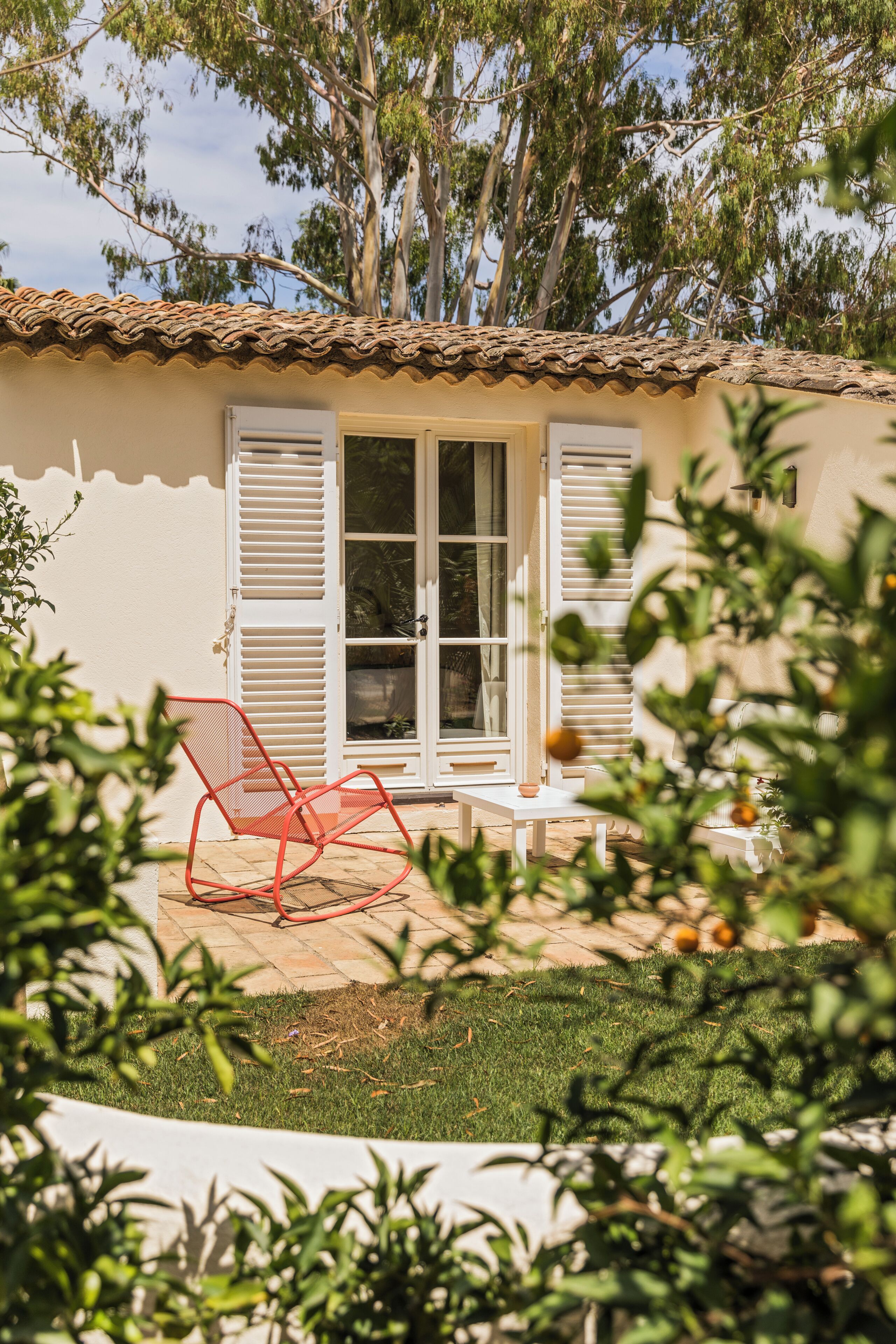Una casa con techo de tejas, puerta blanca con contraventanas y una silla roja al frente.