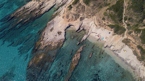 An aerial view of a coastal area with clear turquoise waters, rocky terrain, and a sandy beach with people and beach equipment.