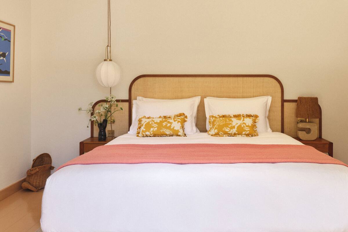 Een opgemaakt bed met witte en gele kussens, een roze deken en een houten hoofdbord.