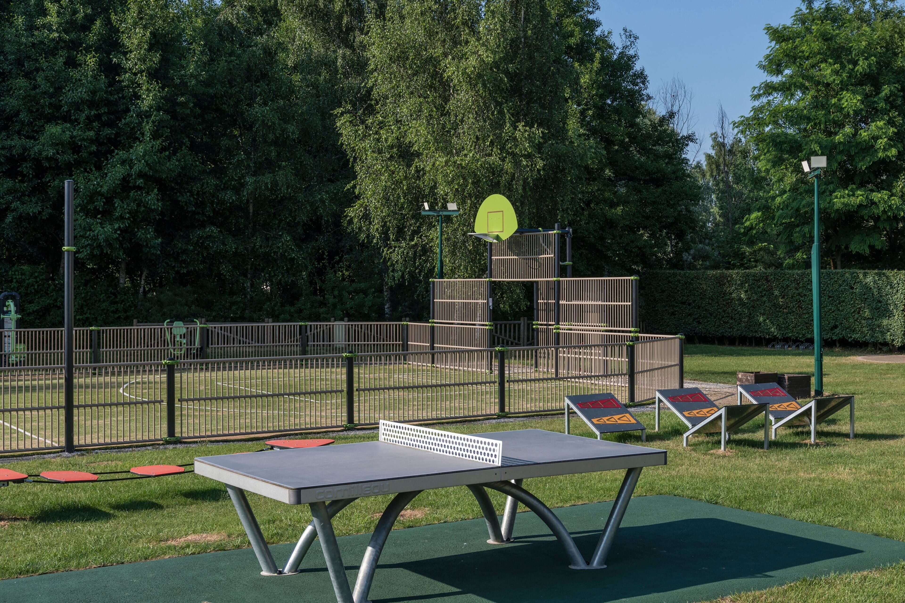 A ping pong table in a park with a fence, trees, and a basketball hoop.