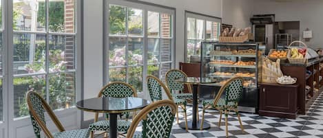 Un café con sillas y mesas con diseños, una vitrina con productos de repostería y ventanas amplias con vista a zonas verdes.