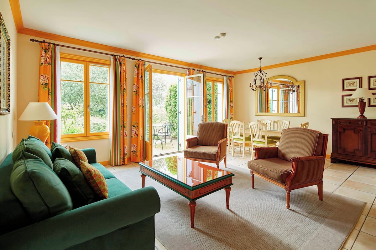 A spacious living room with a green sofa, a wooden chair, a glass coffee table, and a dining area with chairs and a table.