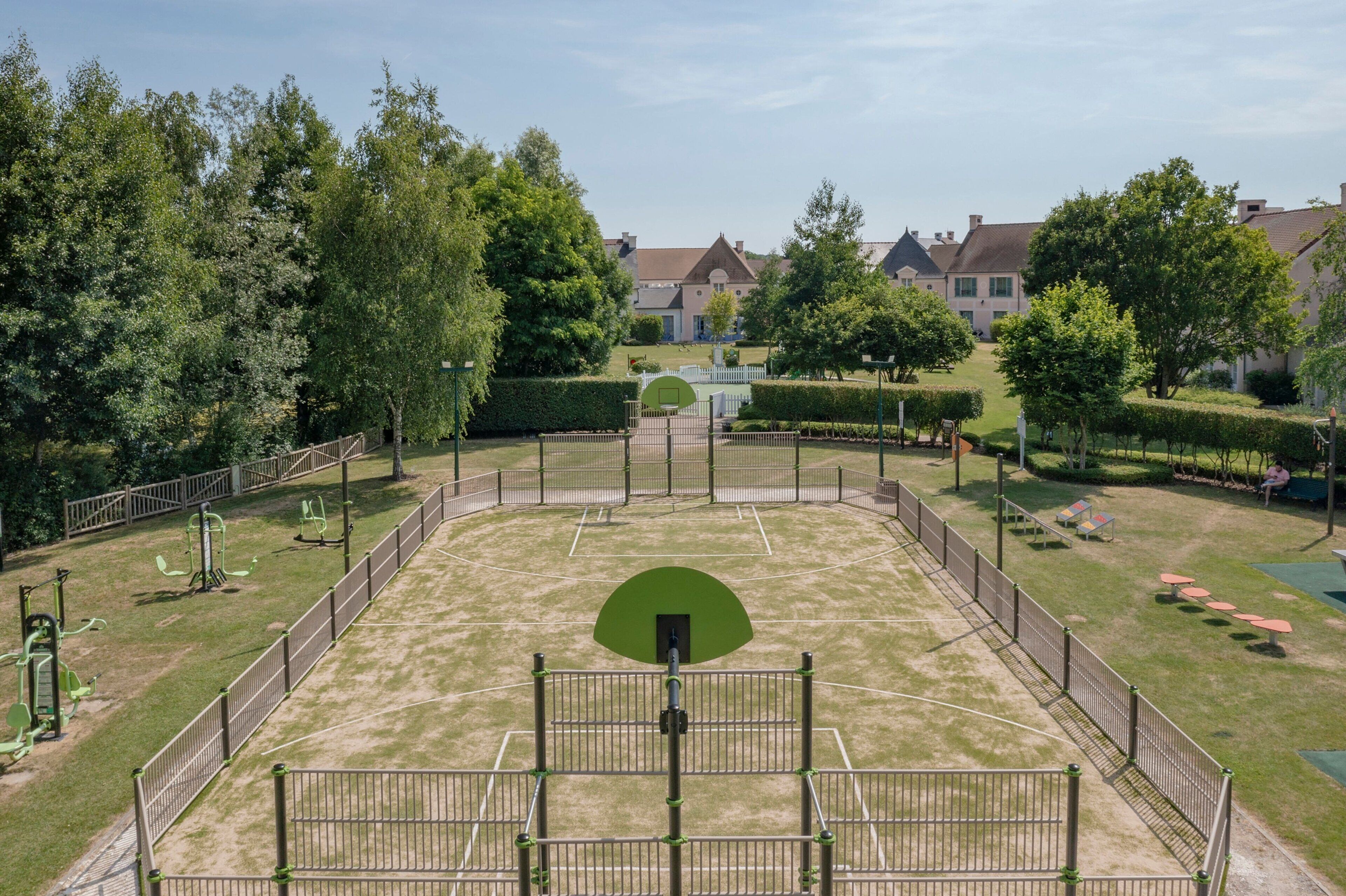 An outdoor basketball court surrounded by a fence, with trees and a building in the background.