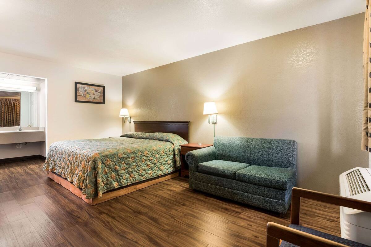 A hotel room with a bed, a green armchair, a wooden chair, a nightstand, a lamp, and a window with curtains.