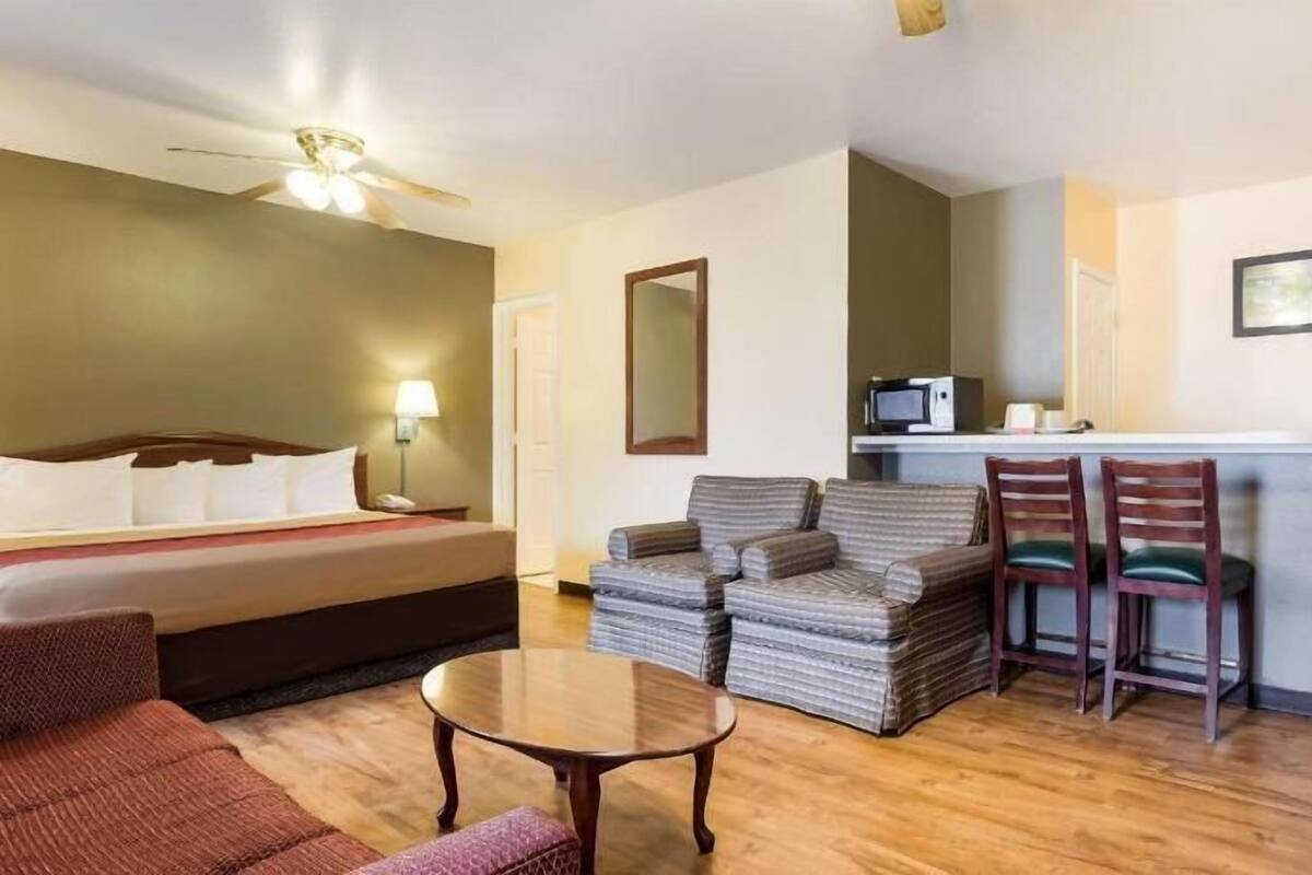 A hotel room with a bed, a desk with a computer, two chairs, a small table, and a ceiling fan.