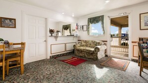 A room with a wooden dining table, a floral-patterned sofa, a red and blue doormat, and a view through a glass door.