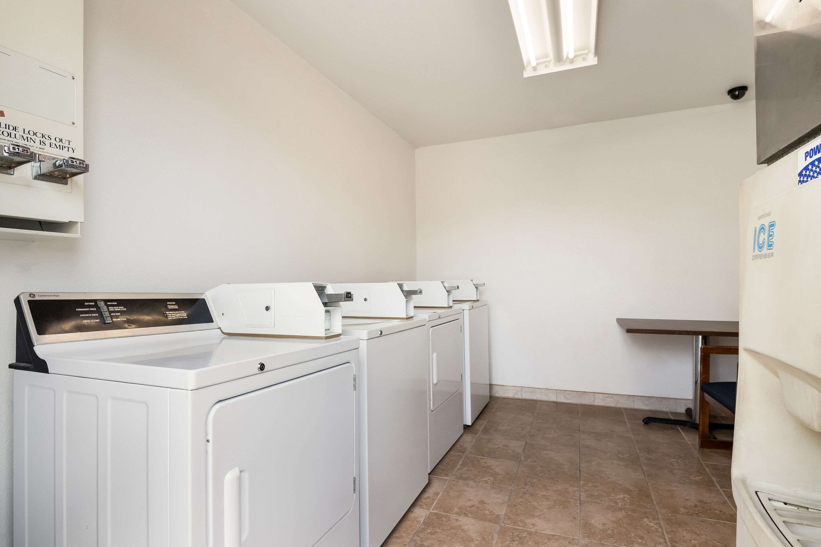 A laundry room with four white front-loading washers and dryers, a control panel, and a small table.