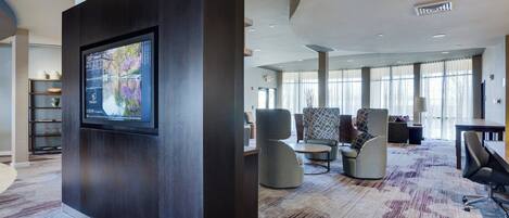 Un moderno vestíbulo de hotel con un televisor de pantalla plana grande montado sobre un panel oscuro, zonas de estar cómodas con sillones y sofás, y un suelo alfombrado.