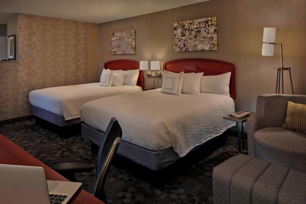 A hotel room with two beds, a desk with a laptop, a chair, and a standing lamp.