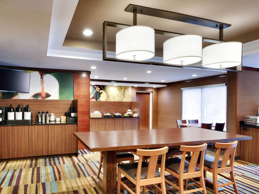 A modern hotel room with a wooden dining table, chairs, a flat-screen TV, a kitchen area, and a large window.