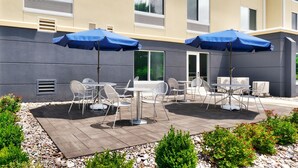 An outdoor patio with tables and chairs, blue umbrellas, and air conditioning units on the building facade. - Fairfield Inn by Marriott Lexington Park Patuxent River Naval Air Station (Lexington Park)