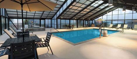 An indoor swimming pool with a glass ceiling, lounge chairs, and a table with chairs.